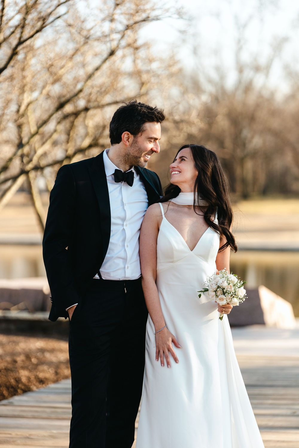 Bride and groom smiling at each other near a serene pond, she in a white gown holding a bouquet, he in a black tuxedo.
