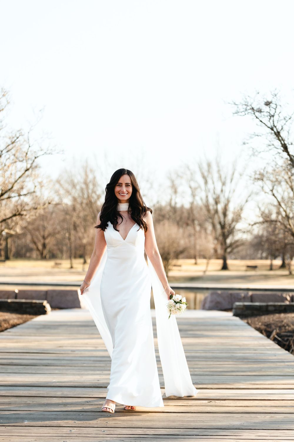 Elegant bride in a sleek white gown holds a bouquet while walking on a wooden bridge in a winter park.