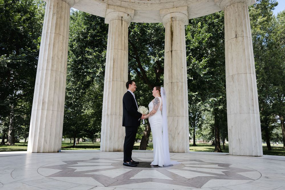 Bride and groom stand facing each other under white marble columns in an outdoor wedding ceremony setting.
