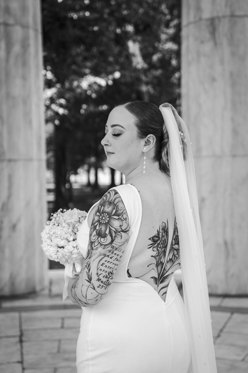 Tattooed bride in backless white gown holds bouquet by marble columns.