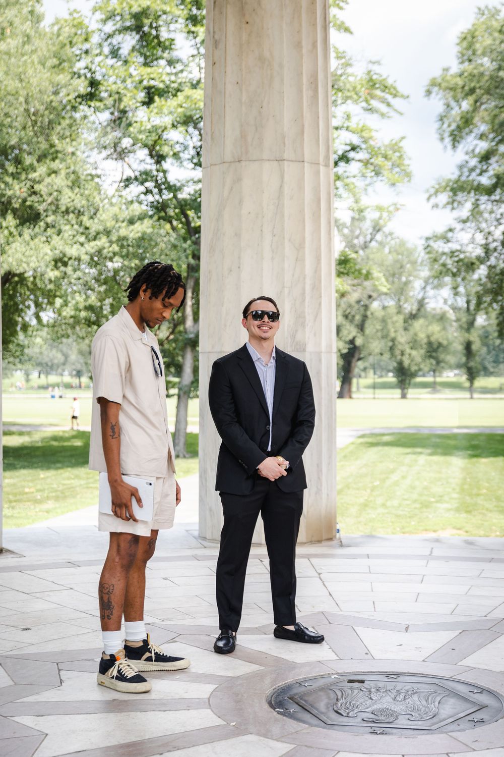 Two men stand between marble columns at an outdoor monument, one in casual wear and one in a black suit.