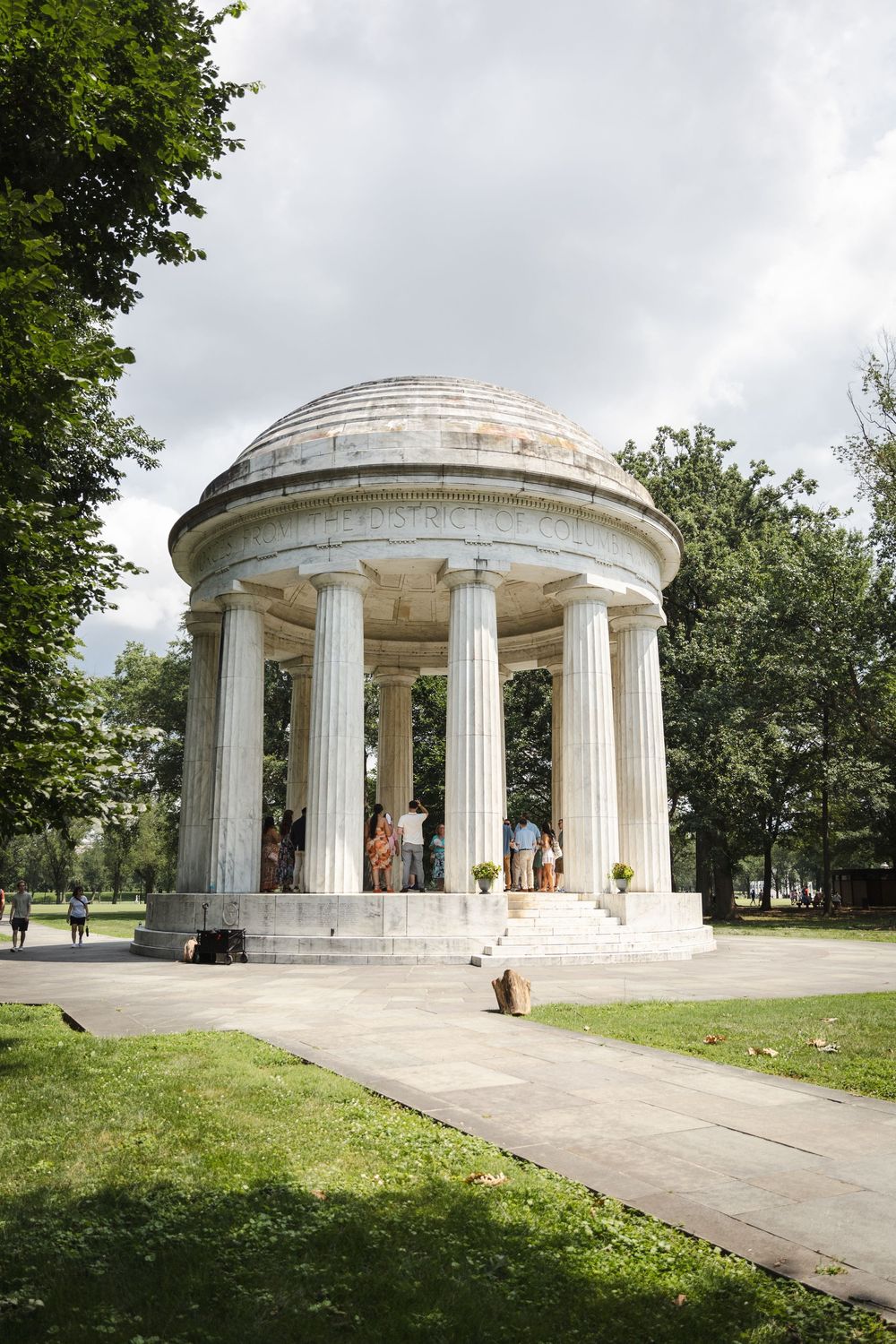 A neoclassical domed marble rotunda with Ionic columns in a lush green park under cloudy skies.