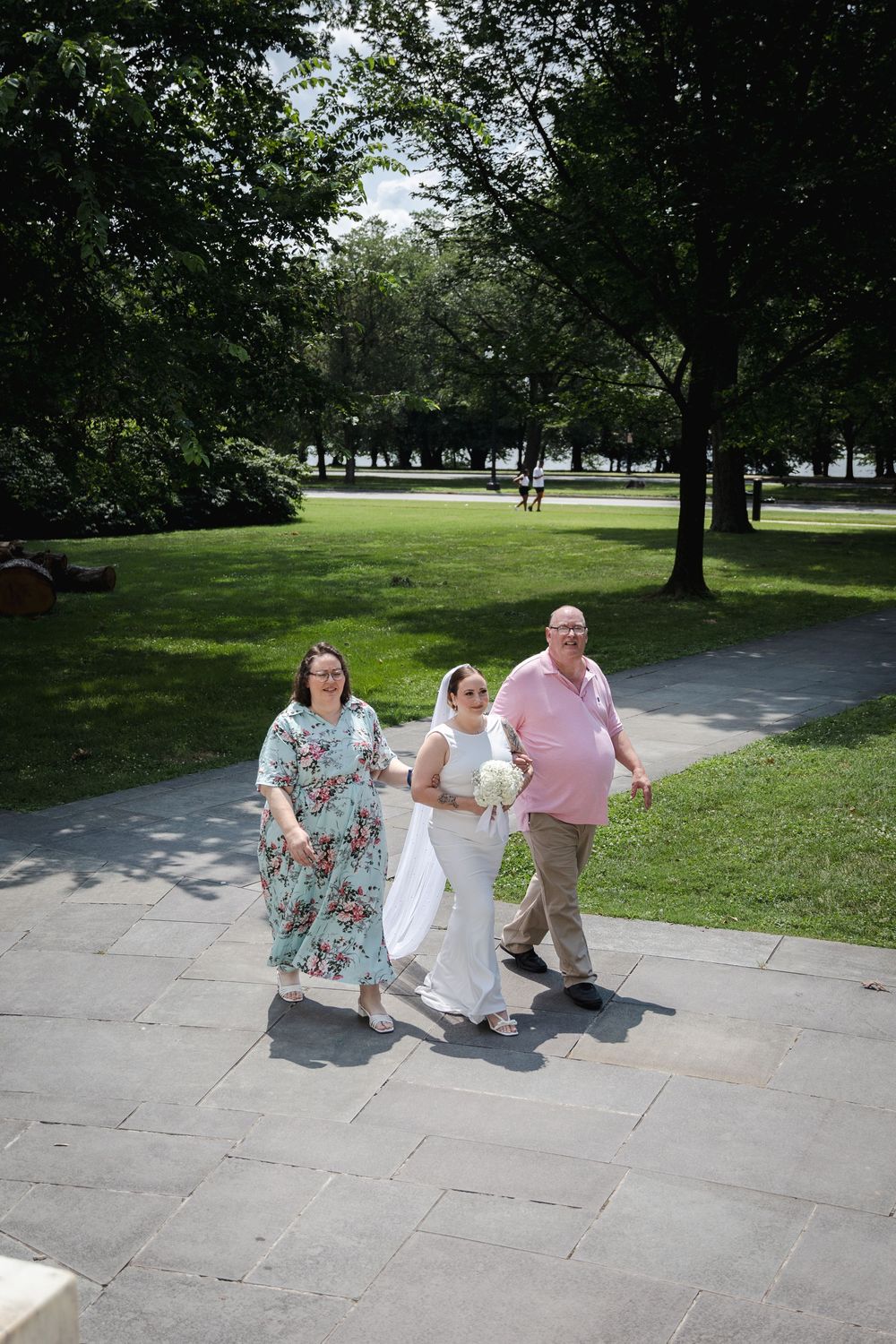 Bride in white gown walks with parents through a sunny park on her wedding day.