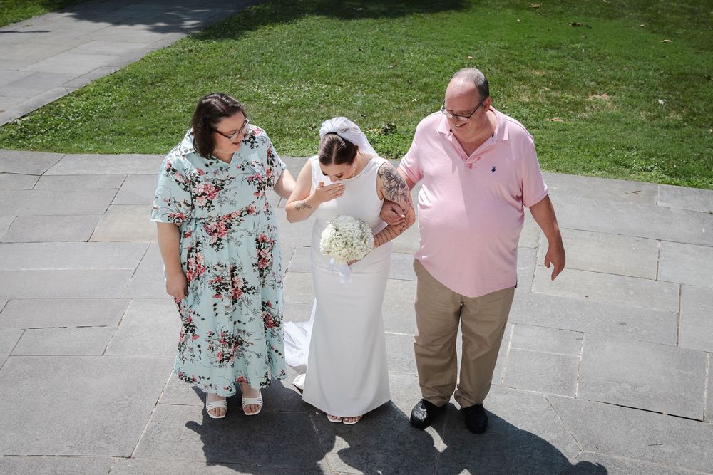 Bride in white gown walks with two guests on stone steps outdoors during a sunny wedding celebration.