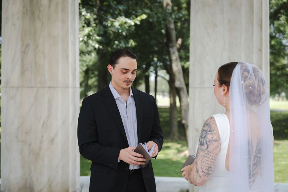 Groom in black suit reads vows to tattooed bride in white dress and veil during outdoor wedding ceremony.