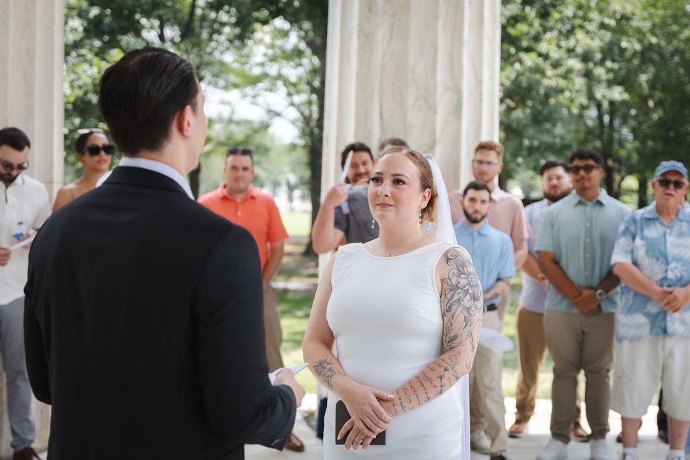 Bride in white dress with tattoo sleeve exchanges vows with groom outdoors at wedding ceremony with guests watching.