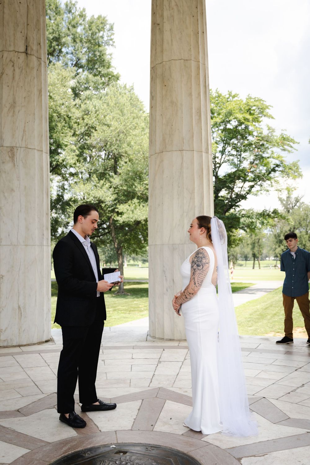 Groom reads vows to bride in white dress and veil at DC memorial columns outdoor wedding ceremony.