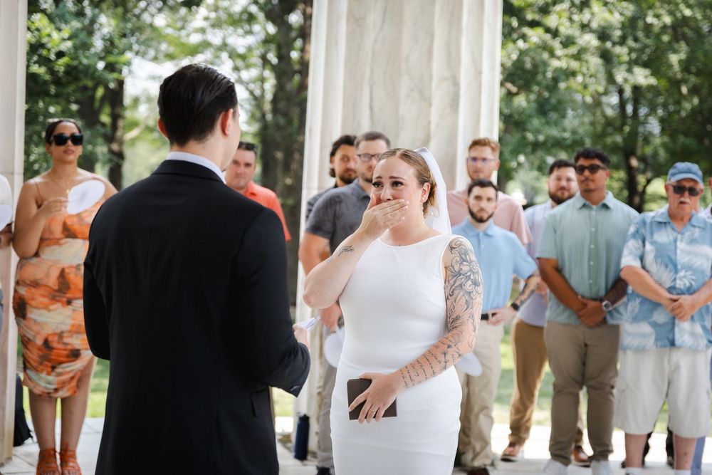 Emotional bride in white sleeveless dress with tattoos covers mouth during outdoor wedding ceremony with guests standing behind.
