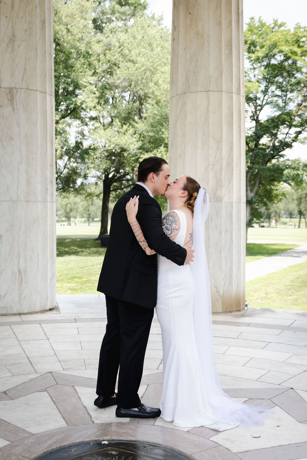 Bride and groom share first kiss in white wedding dress and black tuxedo between marble columns.