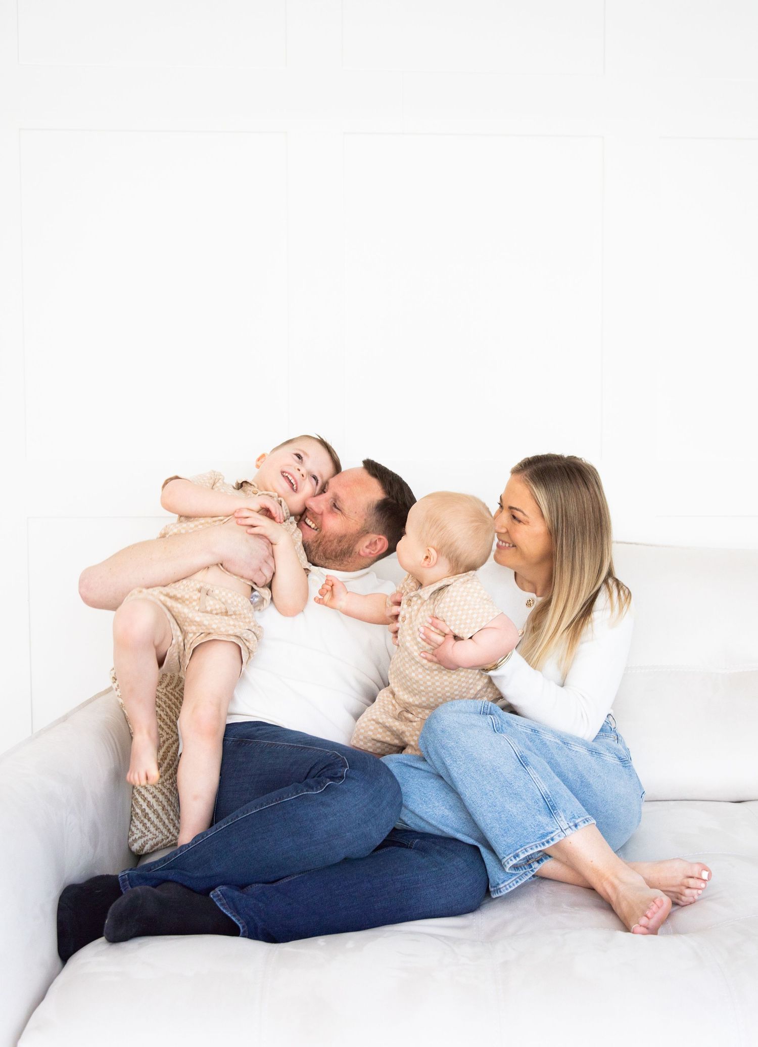 A happy family of four sits on a white couch, laughing and playing with their toddler and baby.