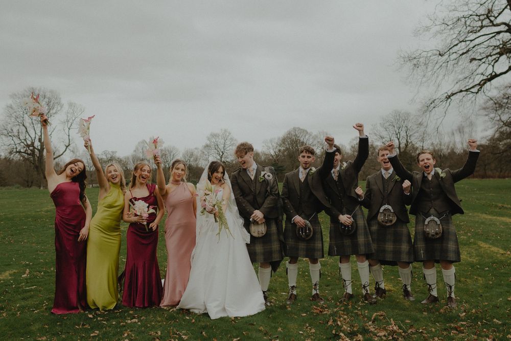 Wedding party celebrating outdoors in Scotland, bridesmaids in colorful gowns and groomsmen in kilts cheering on a green lawn.
