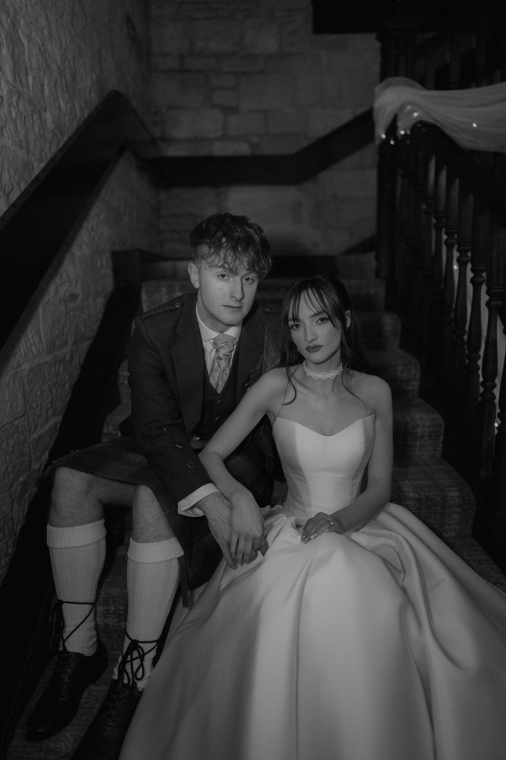 Black and white photo of a couple in wedding attire sitting on stone stairs, bride in ball gown, groom in suit.