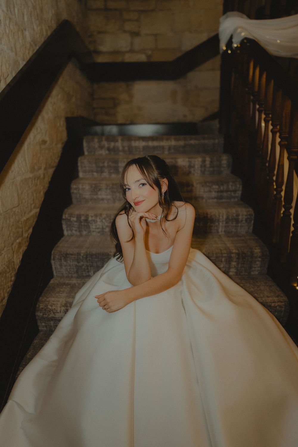 Bride in a white ball gown sitting gracefully on stone stairs with dark wood railings.