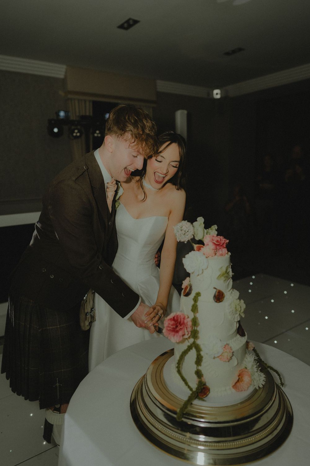 Bride and groom laughing while cutting a three-tier white wedding cake decorated with pink and white flowers.