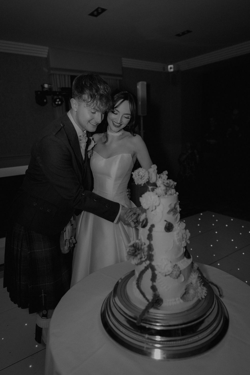 Black and white photo of a bride and groom cutting their floral wedding cake, groom wearing a kilt.