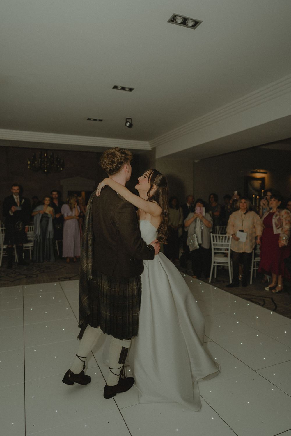 Bride and groom sharing their first dance on a white LED dance floor surrounded by wedding guests.