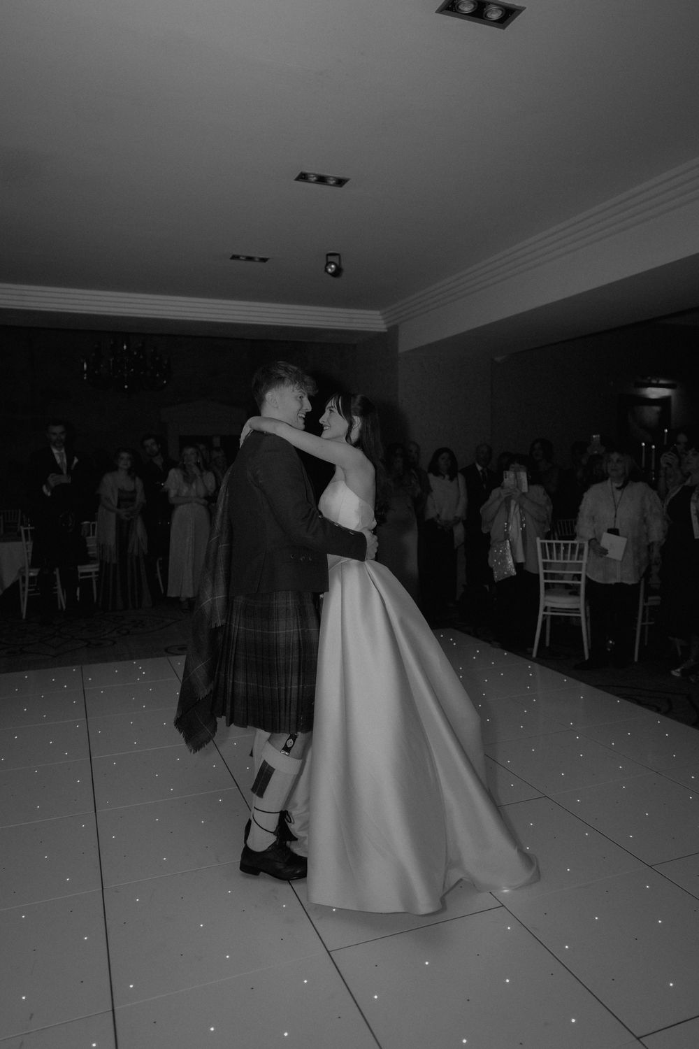 Black and white photo of a bride and groom sharing their first dance at a wedding reception with guests watching.