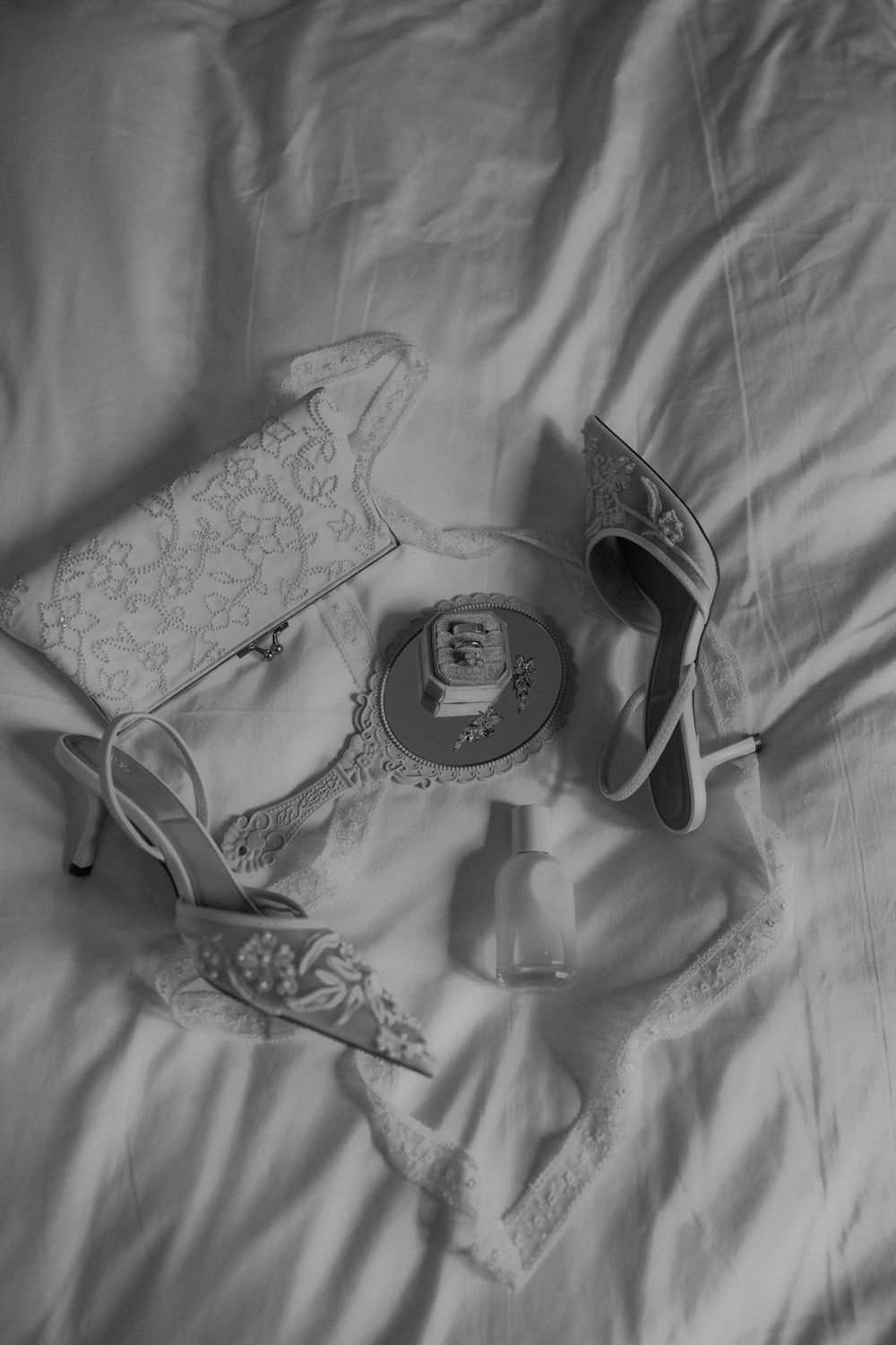 Black and white flat lay of bridal accessories including lace heels, clutch purse, perfume bottle, and compact mirror on white bedding.