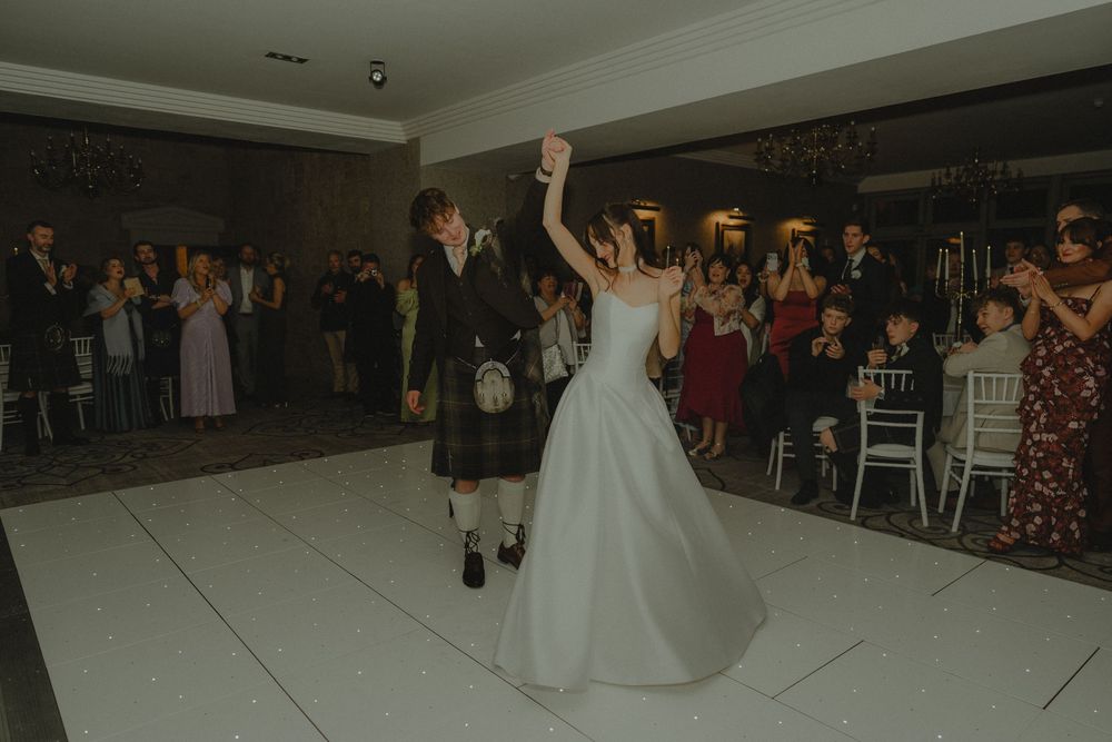 Bride and groom share their first dance at a wedding reception, surrounded by guests on a white dance floor.