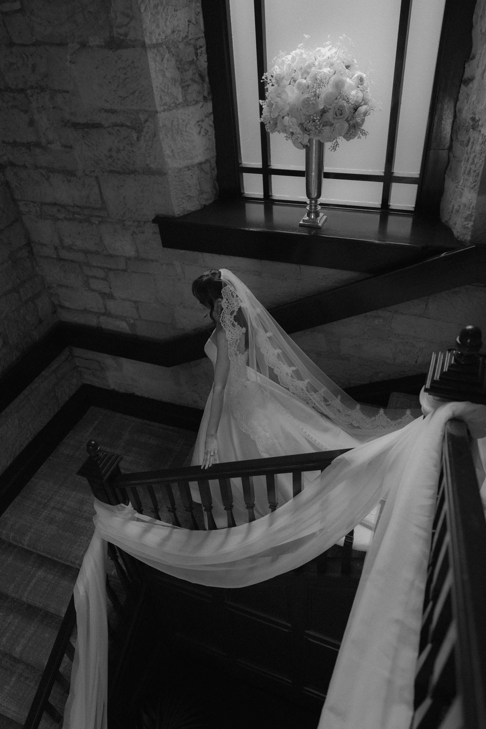 Black and white photo of a bride in a flowing wedding gown and veil descending an elegant staircase with floral arrangement.