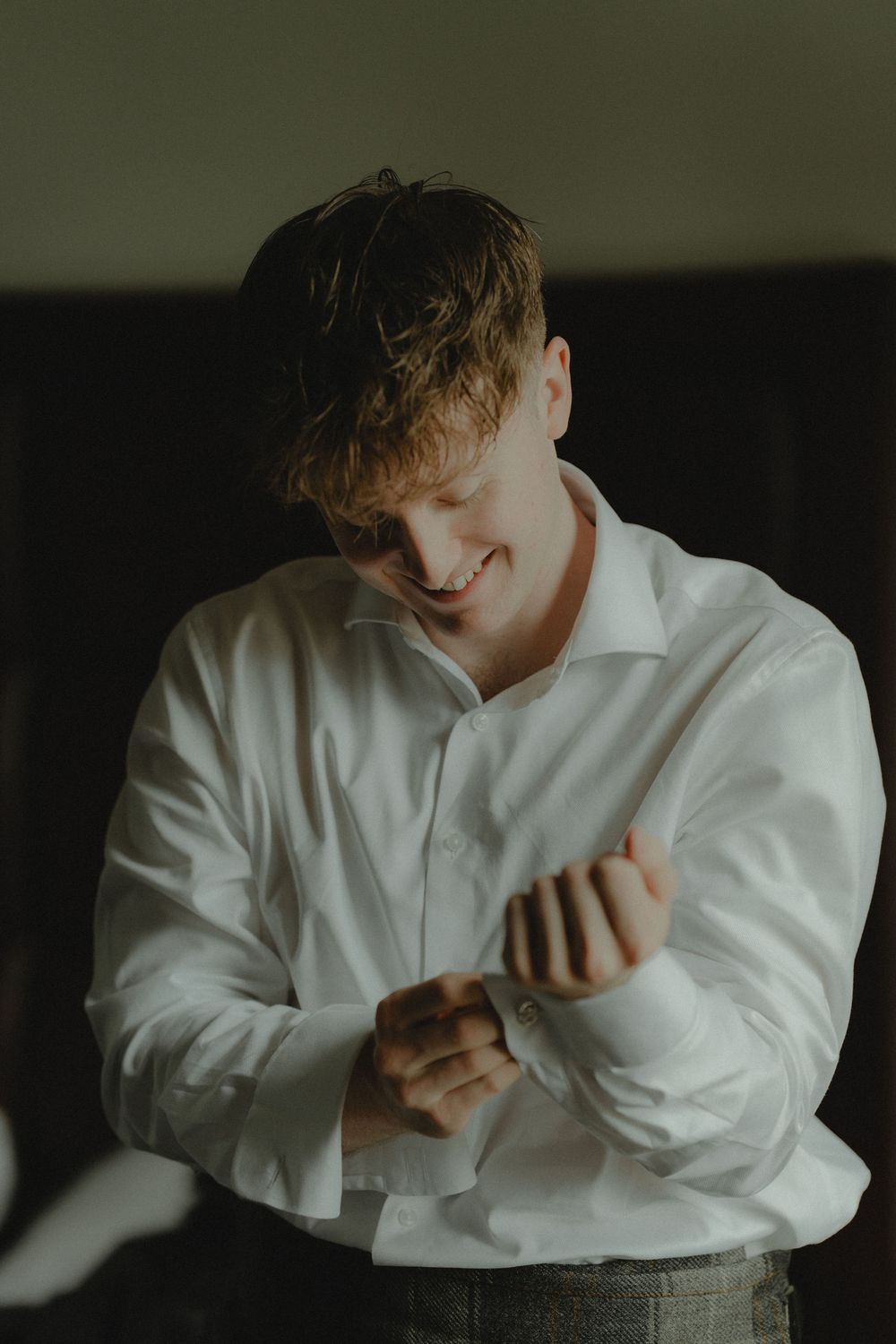 A smiling groom in a white dress shirt fastening his cufflinks while getting ready for his wedding day.