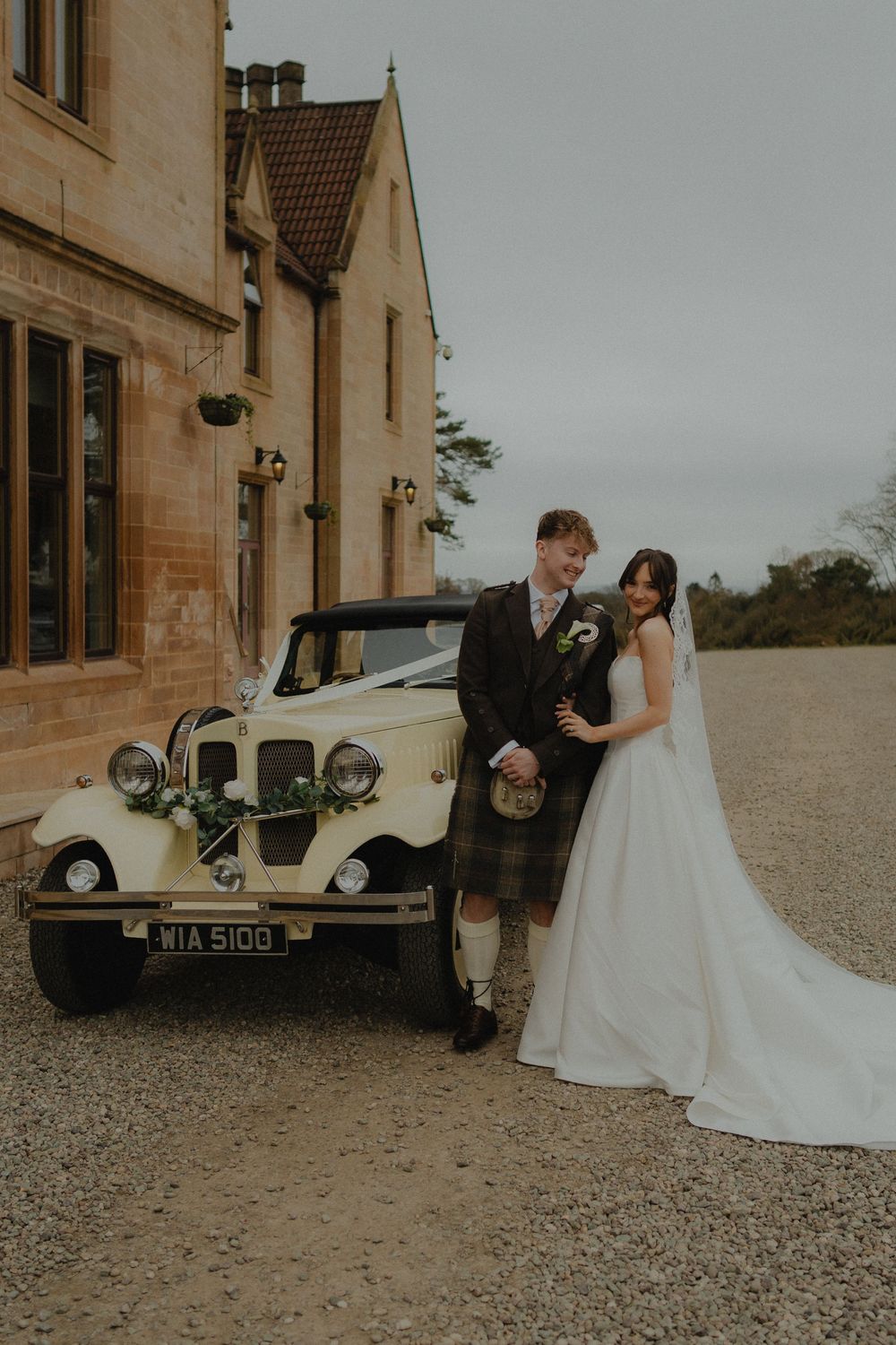 Bride and groom posing beside a vintage cream wedding car outside a stone manor house, groom wearing a tartan kilt.