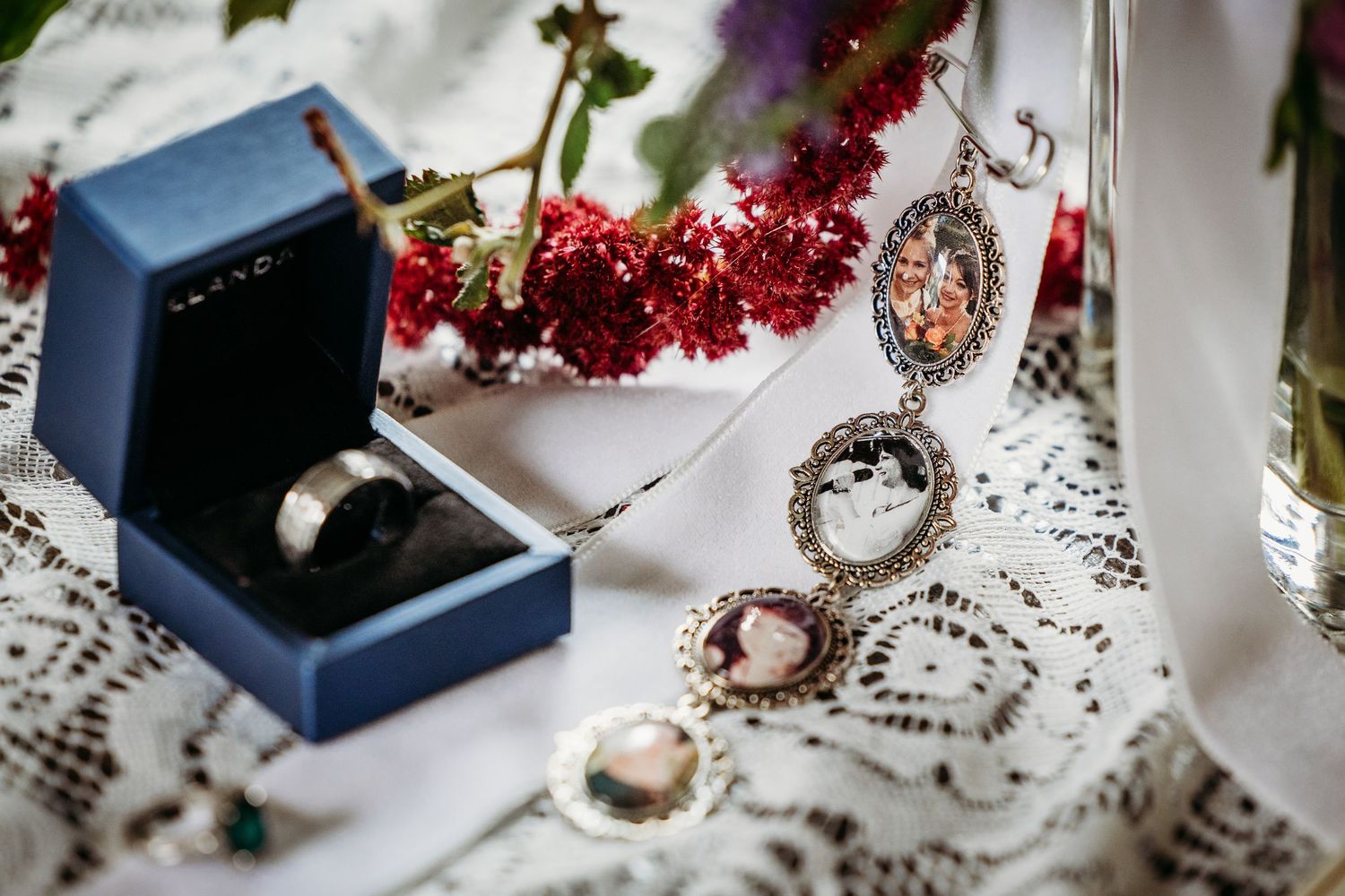 Wedding ring in blue box beside silver locket necklace with photos on white lace fabric in DFW.