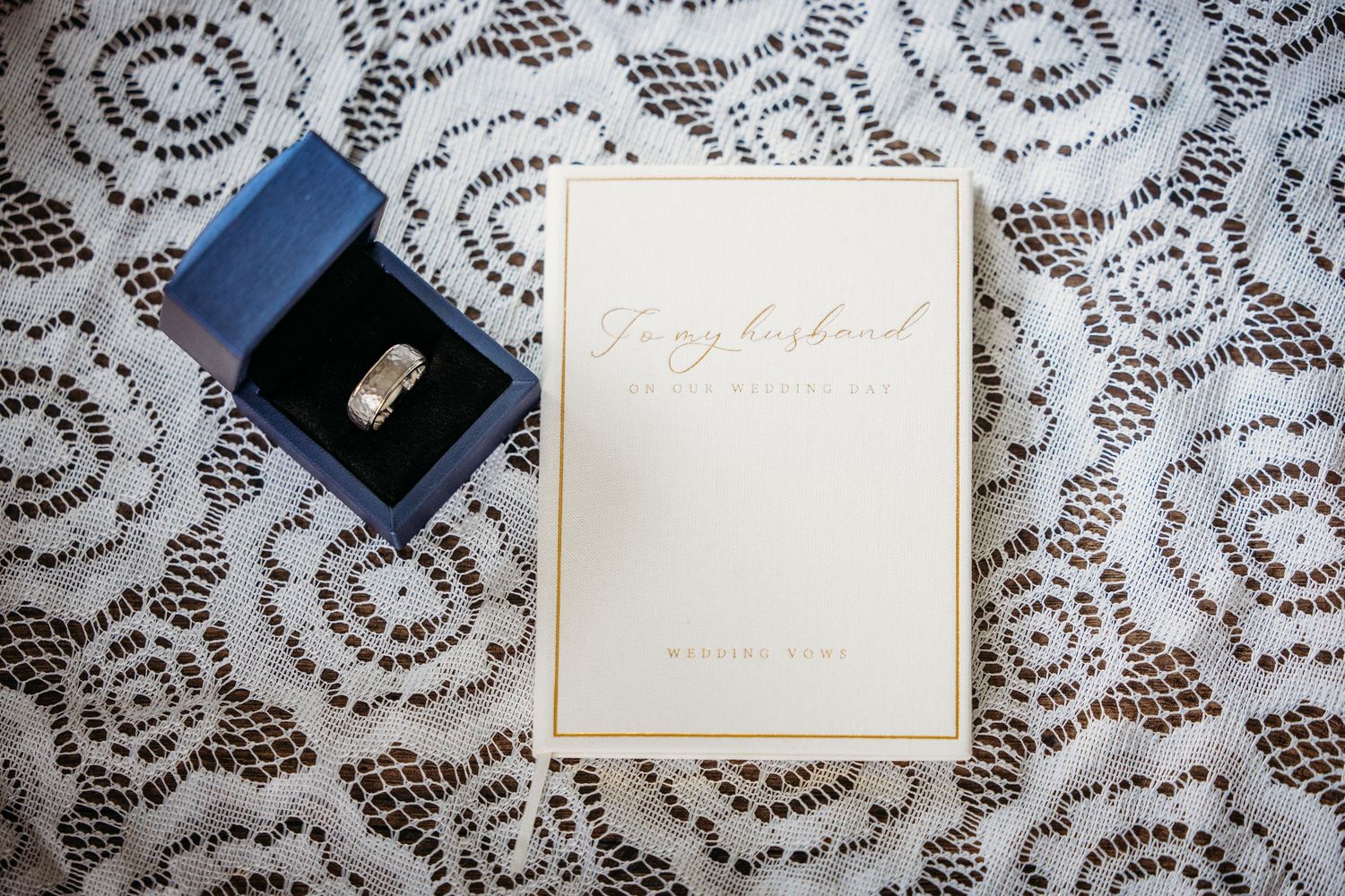 Wedding ring in blue box beside a gold-trimmed card reading To My Husband on white lace fabric in DFW.
