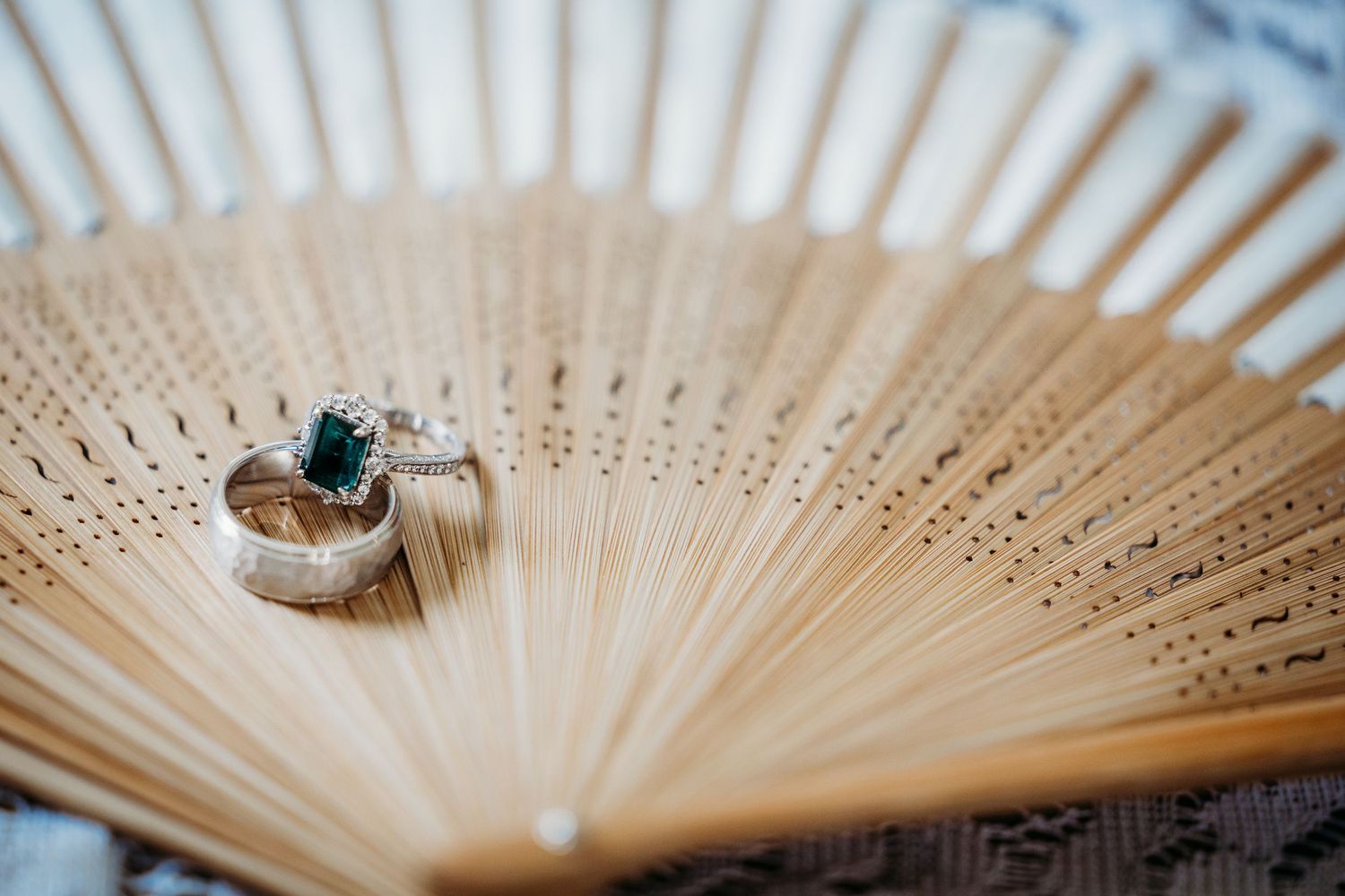Two wedding rings with emerald gemstone displayed on a wooden hand fan in Fort Worth, TX.