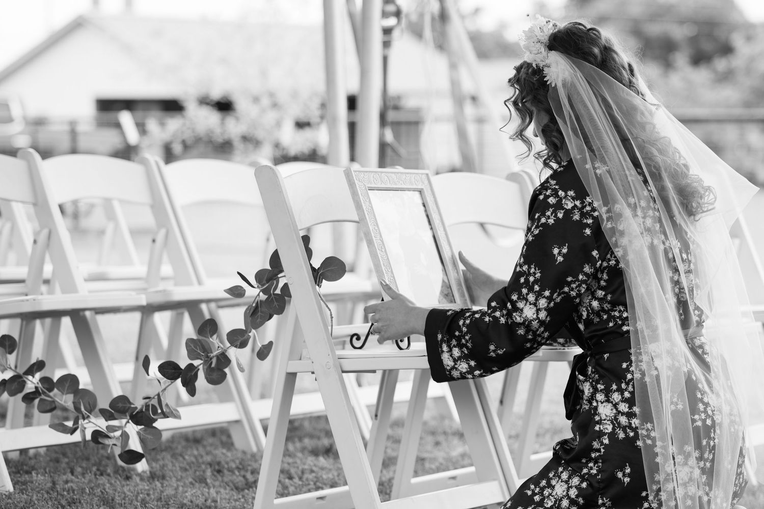 Bride in floral dress and veil arranging a portrait of her mother on a white folding chair at an outdoor wedding ceremony in Fort Worth, TX.