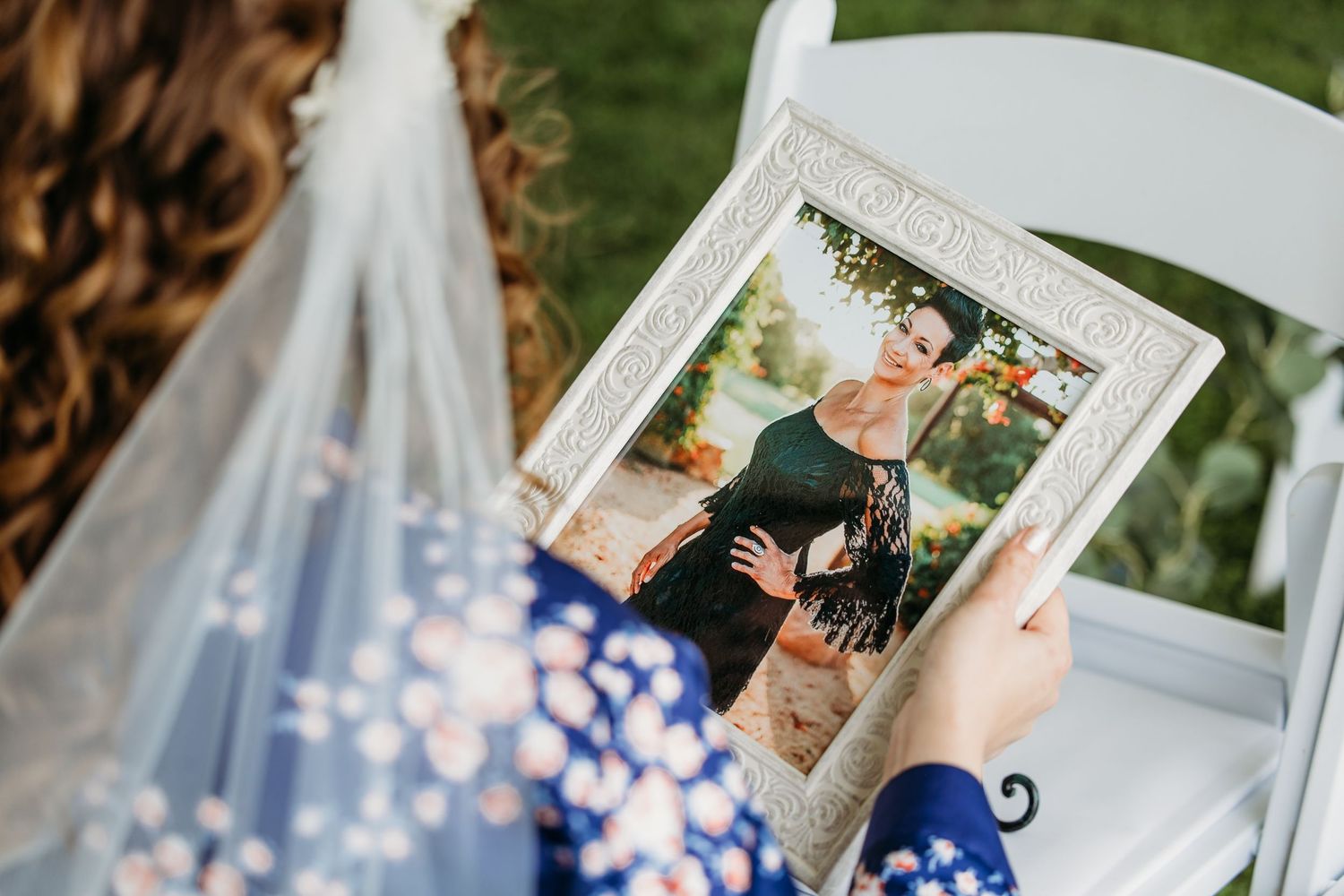 Bride in floral dress and veil arranging a portrait of her mother on a white folding chair at an outdoor wedding ceremony in Fort Worth, TX.