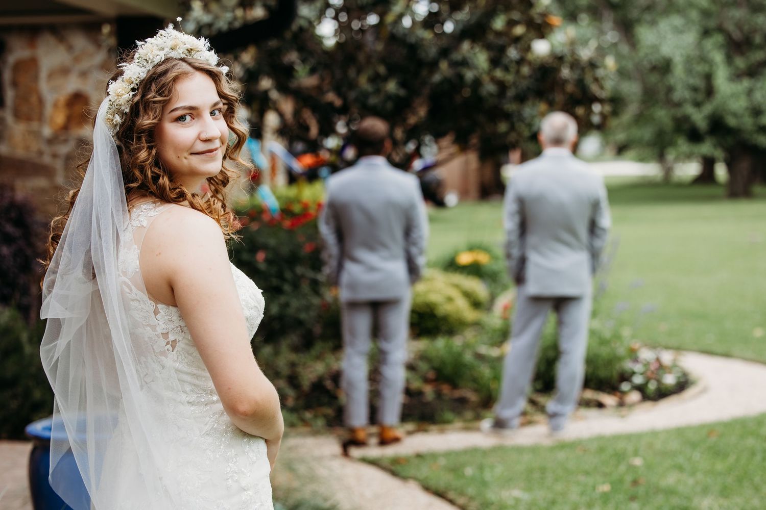 Bride in lace gown and floral crown smiles over shoulder outdoors with two groomsmen in gray suits in background in DFW.