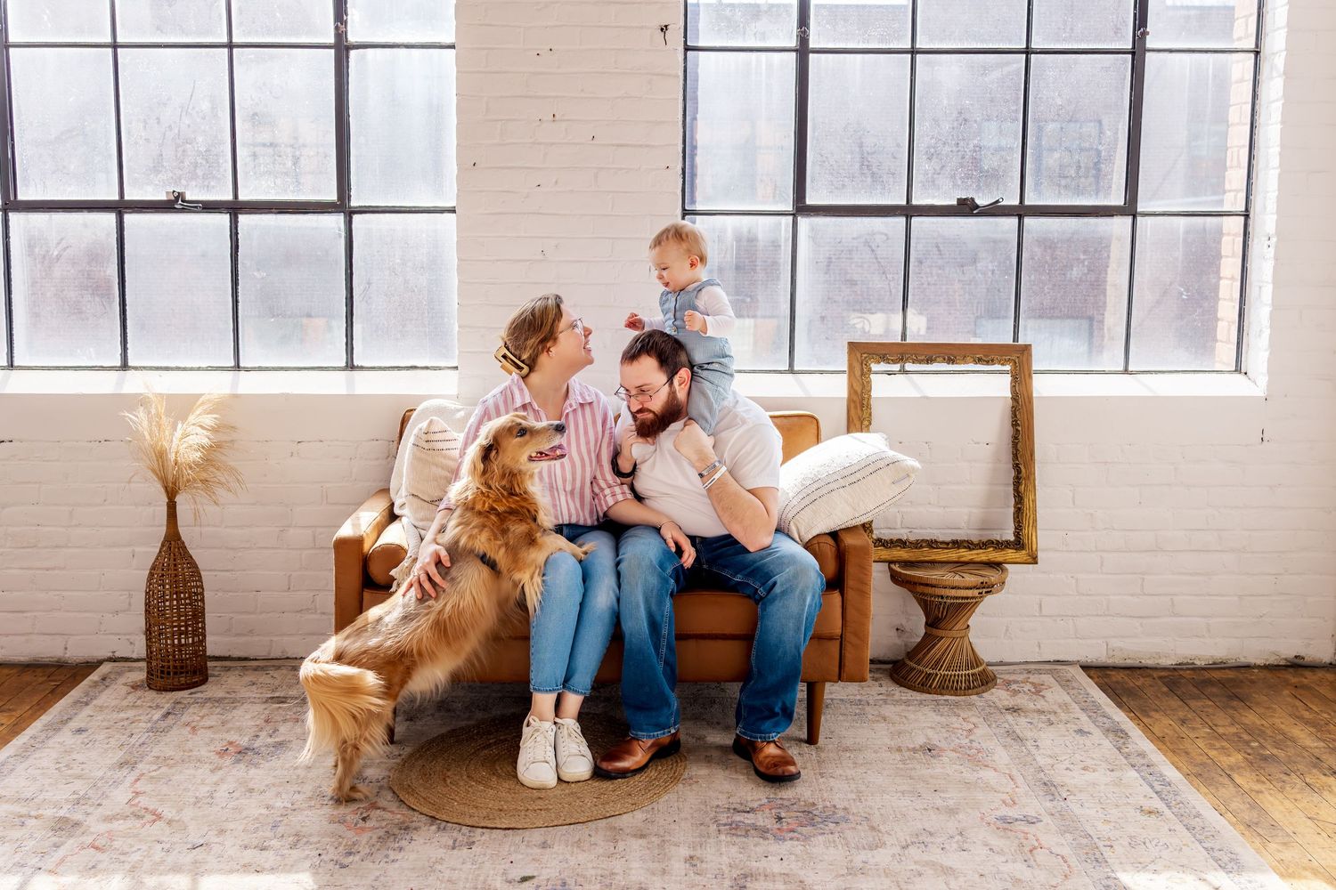Happy family with baby and golden retriever dog sitting together on brown sofa in bright modern living room.