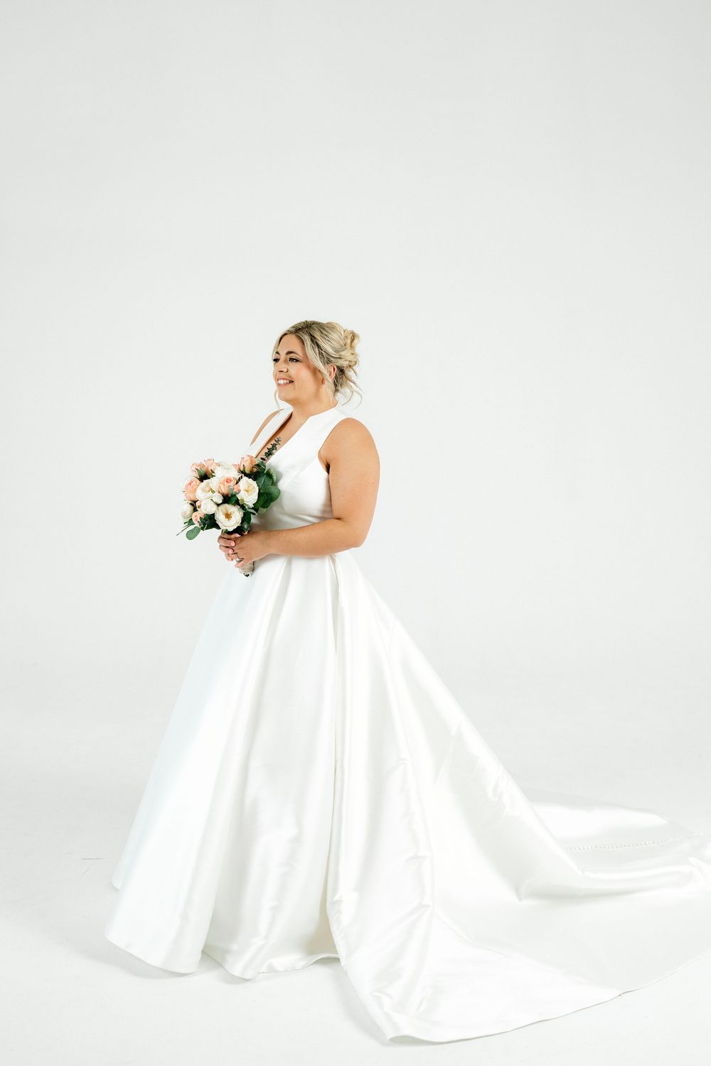 Bride in elegant white ball gown with train holds pink bouquet, posing sideways in studio.