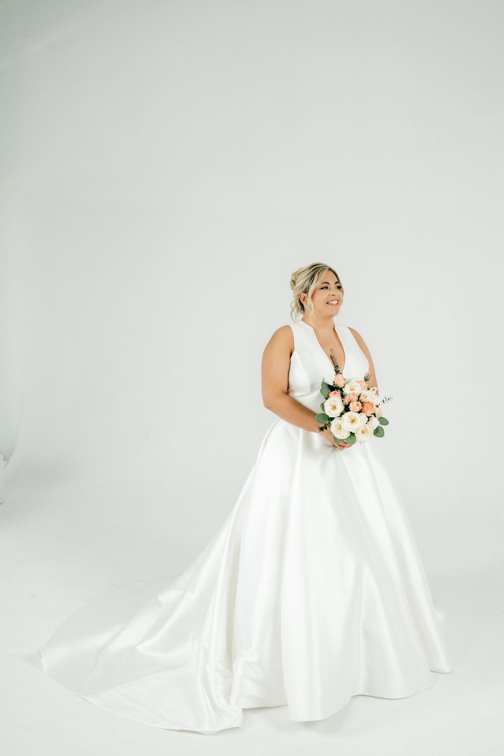 Smiling bride in white deep-V ball gown holds peach and white bouquet in bright studio setting.