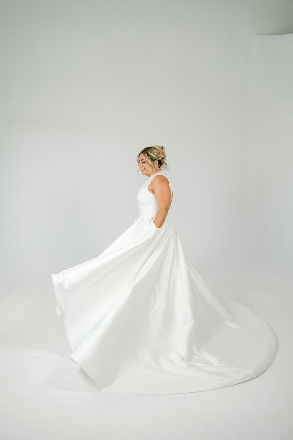 Bride twirling in elegant white satin ball gown with flowing skirt in bright studio setting.