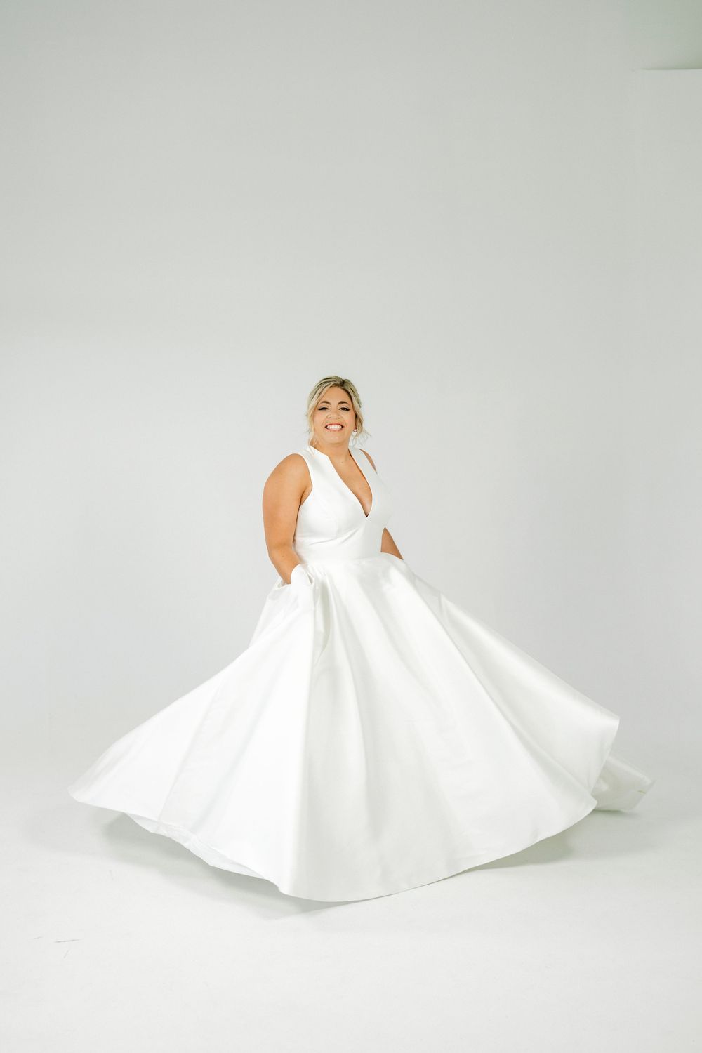 Smiling bride twirling in a white ballgown wedding dress with deep V-neckline and full skirt in a studio setting.