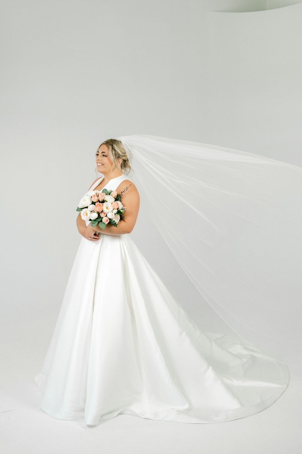 Bride in a white ball gown with flowing veil holds a pink bouquet, smiling in a bright studio setting.