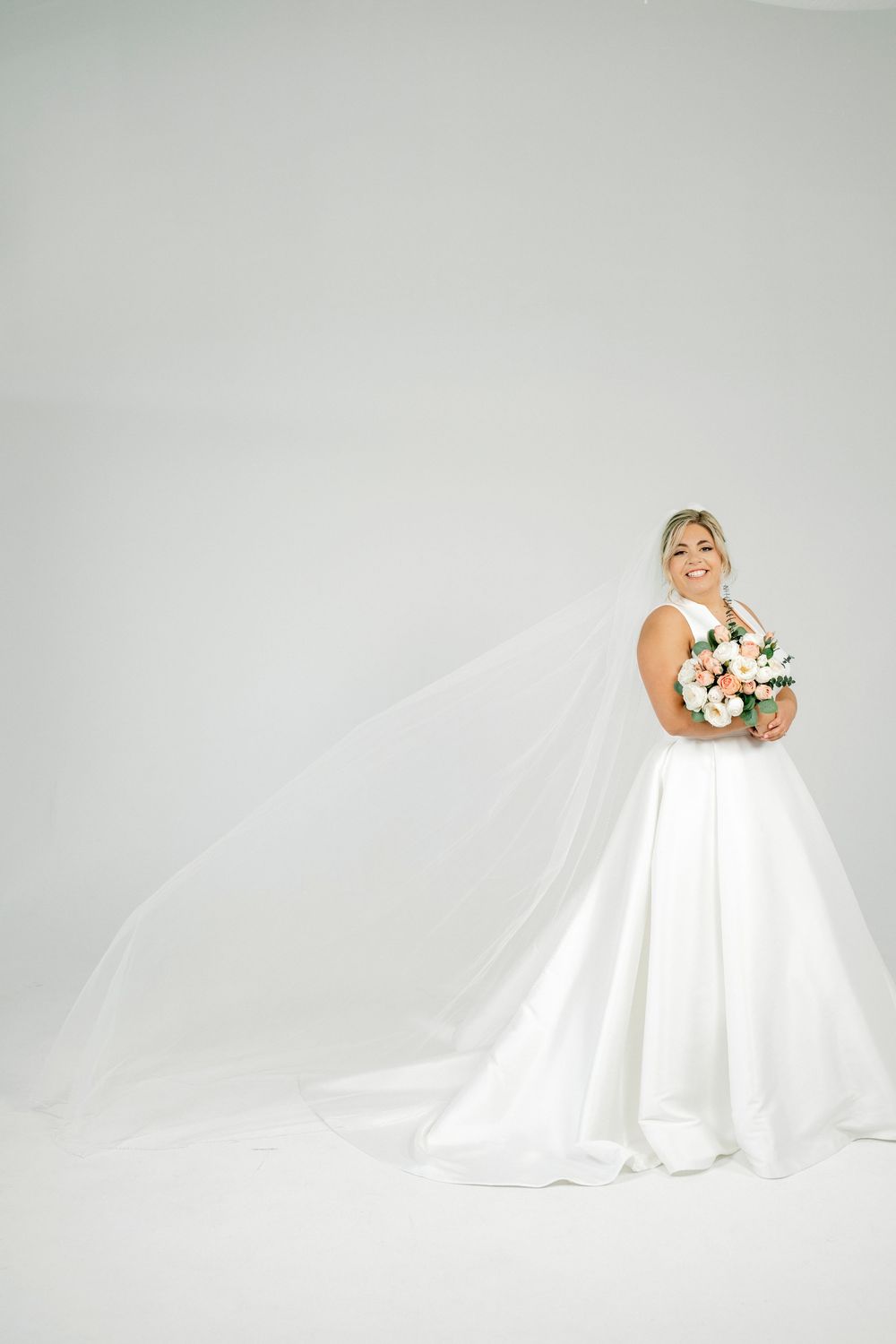 Smiling bride in white ballgown with flowing veil holds pink and white bouquet against light gray background.