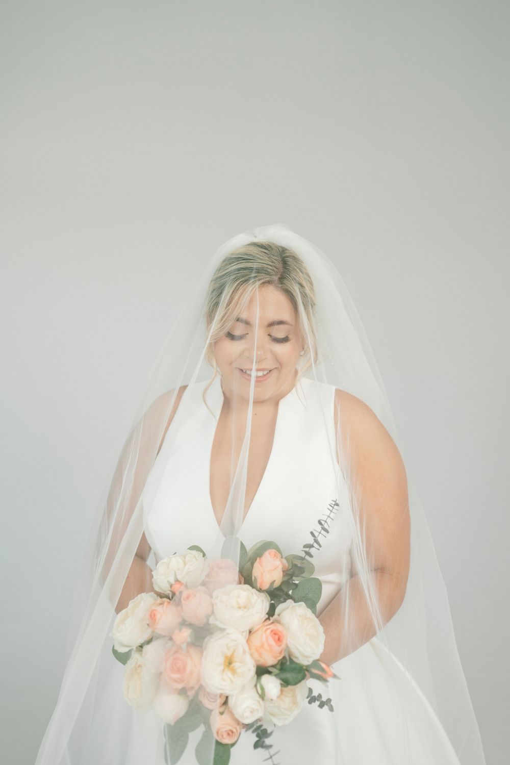 Smiling bride in white sleeveless gown and veil holds a pink and white rose bouquet against a soft gray background.