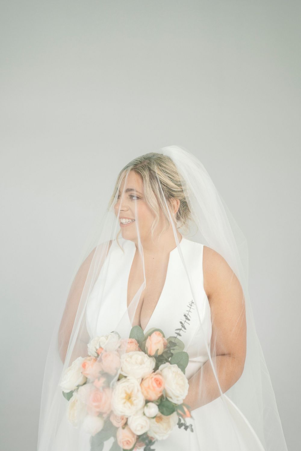Smiling bride in white gown and veil holding peach and white rose bouquet against light gray background.
