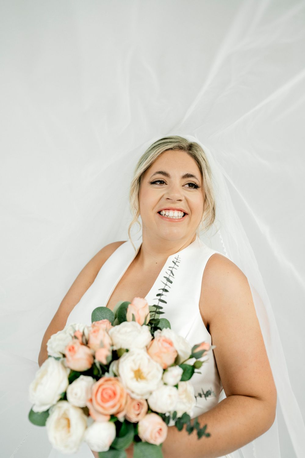 Smiling bride in white sleeveless gown holds pink and white rose bouquet against flowing white fabric backdrop.