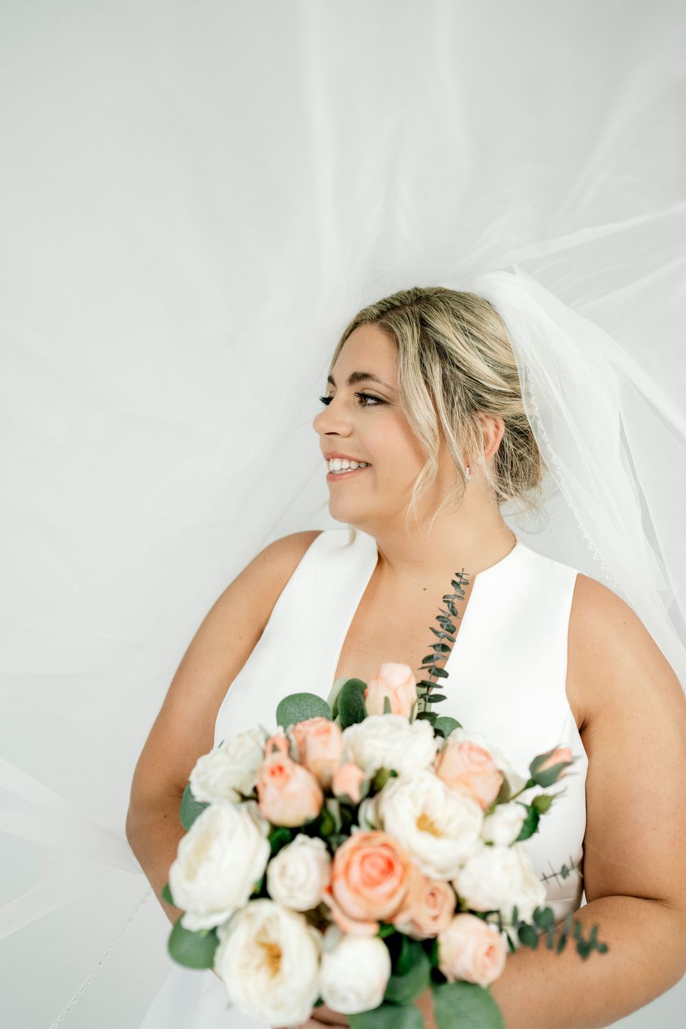 Radiant bride wearing white sleeveless wedding dress and veil holding lush peach and white rose bouquet.