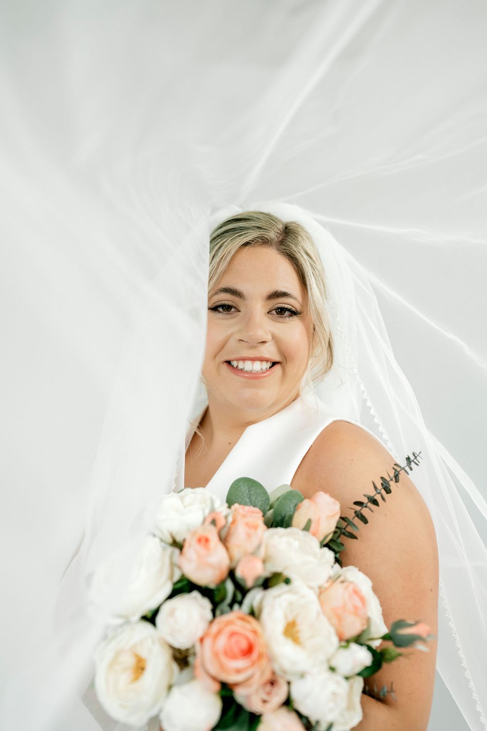 Smiling bride in white dress and veil holding a bouquet of pink and white roses.