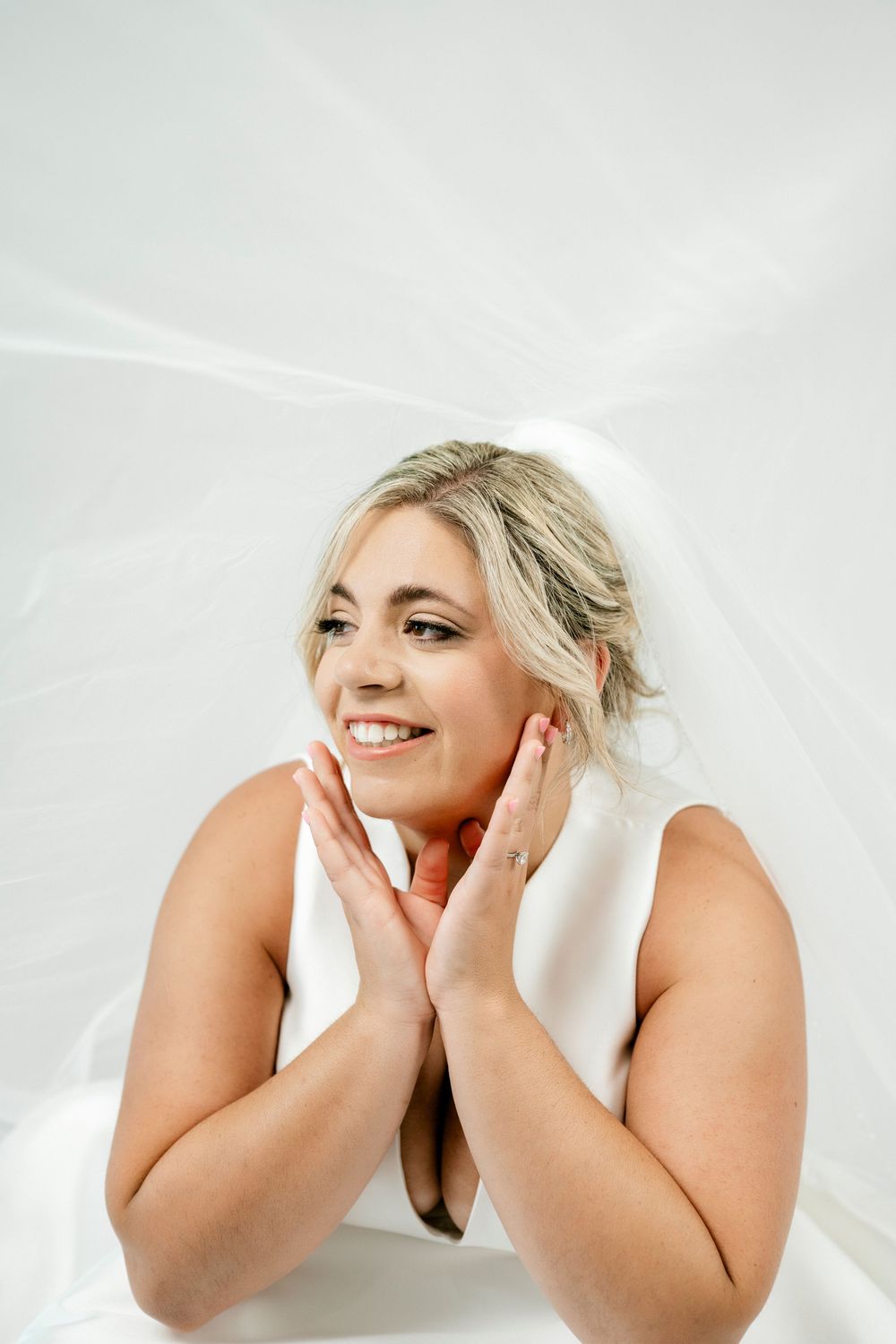 Smiling blonde bride posing under sheer white veil with hands framing her face.