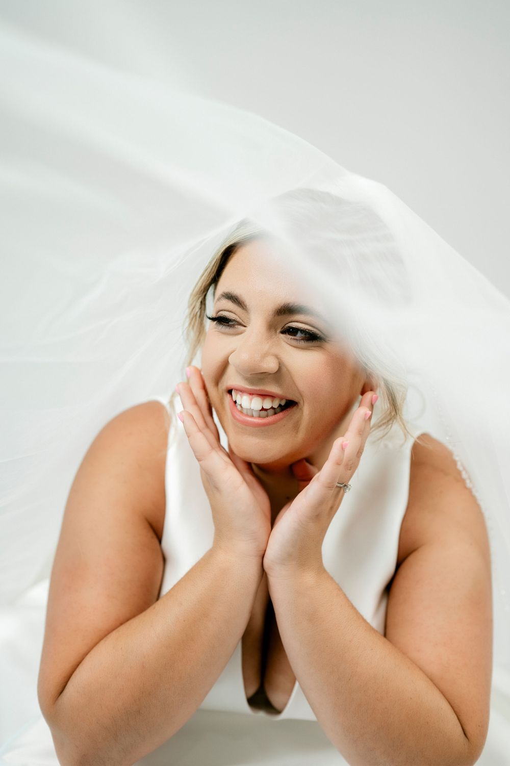Smiling bride laughing under flowing white veil with hands framing face.