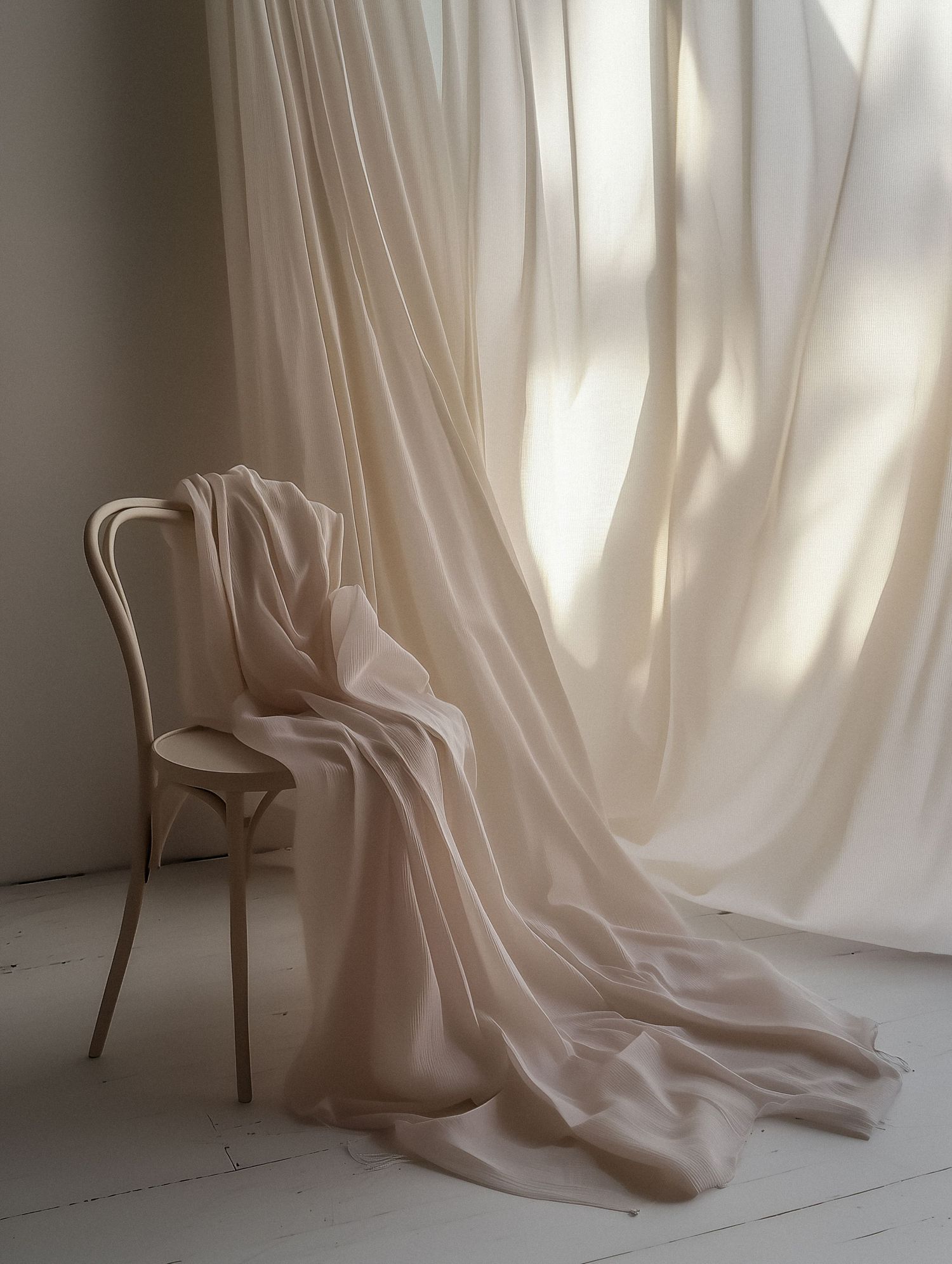 A delicate blush fabric draped over a wooden Thonet chair against sheer white curtains in soft natural light.