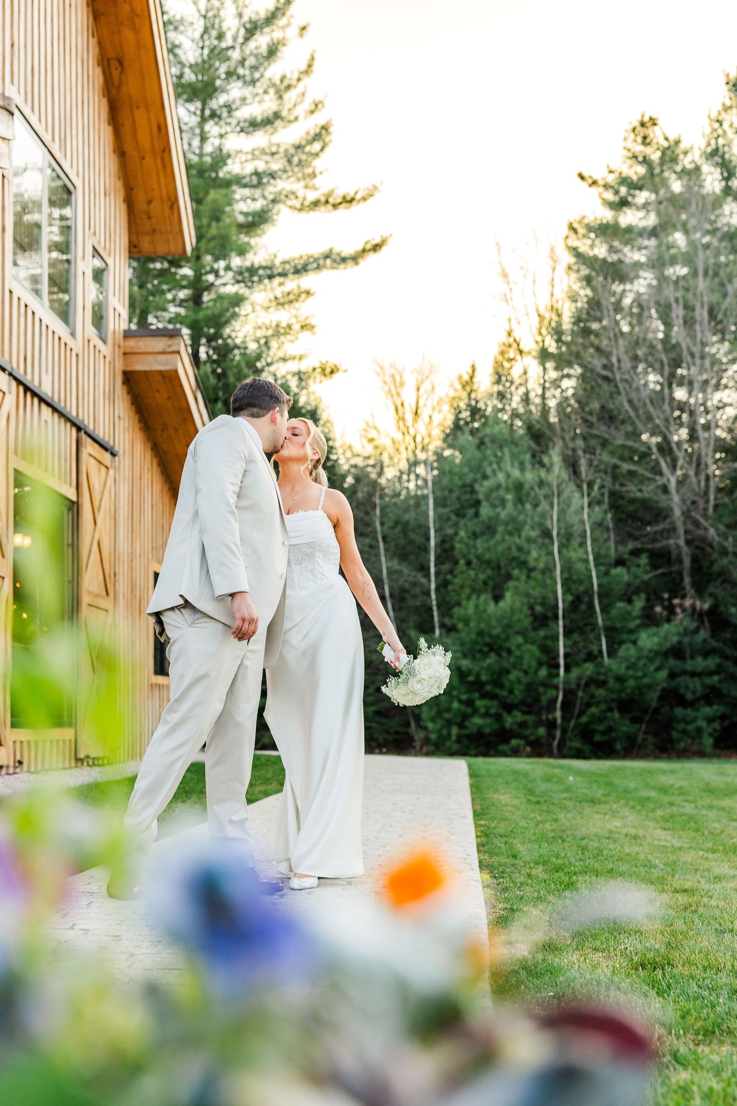 Bride and groom share a romantic moment outside a wooden barn venue surrounded by lush green trees at golden hour.