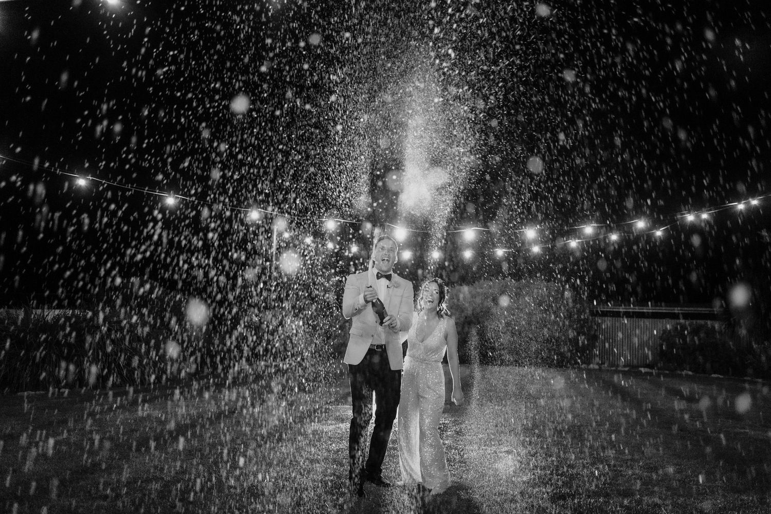 Black and white dramatic photo of couple dancing in heavy rain under string lights at night.