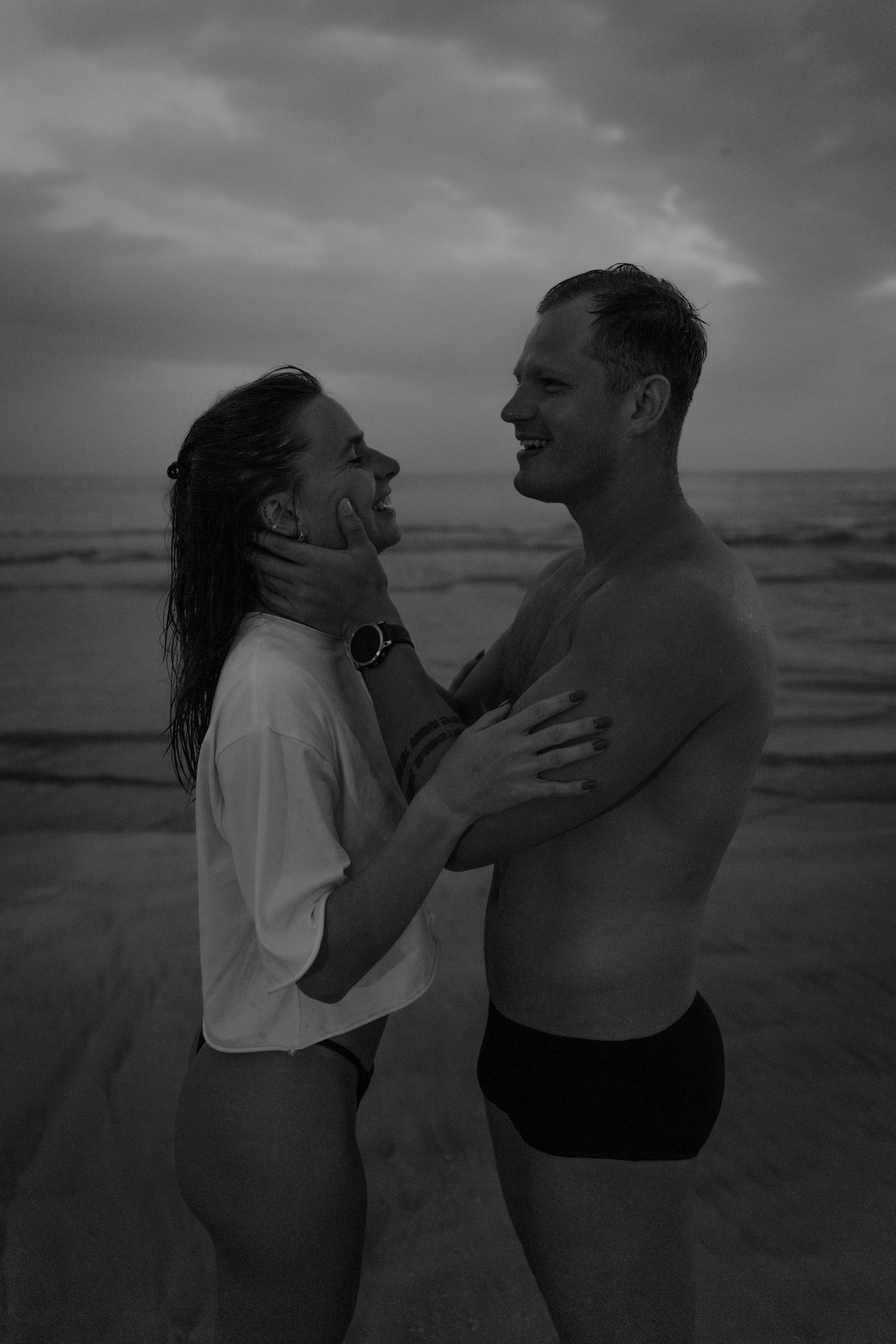 Black and white photo of a couple embracing on a beach, smiling at each other at dusk.